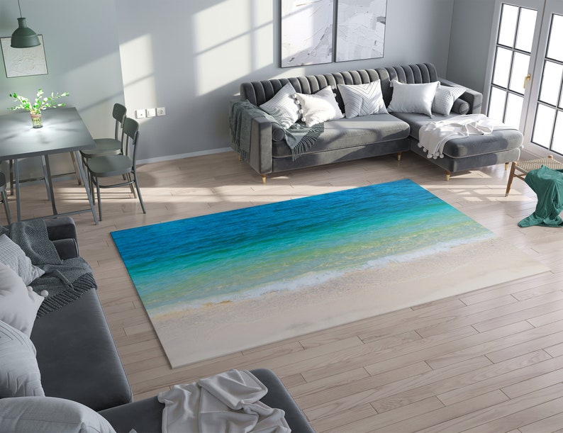 May include: A rectangular area rug featuring a beach scene with turquoise water and white sand. The rug's design mimics the ocean's edge, with varying shades of blue and green transitioning to a sandy white at the bottom. The rug is placed on a hardwood floor.