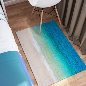 May include: Rectangular rug featuring a beach scene with white sand and turquoise water. The rug's design transitions from a sandy beige to shades of blue and green, creating a coastal aesthetic. The rug is placed on a wooden floor.