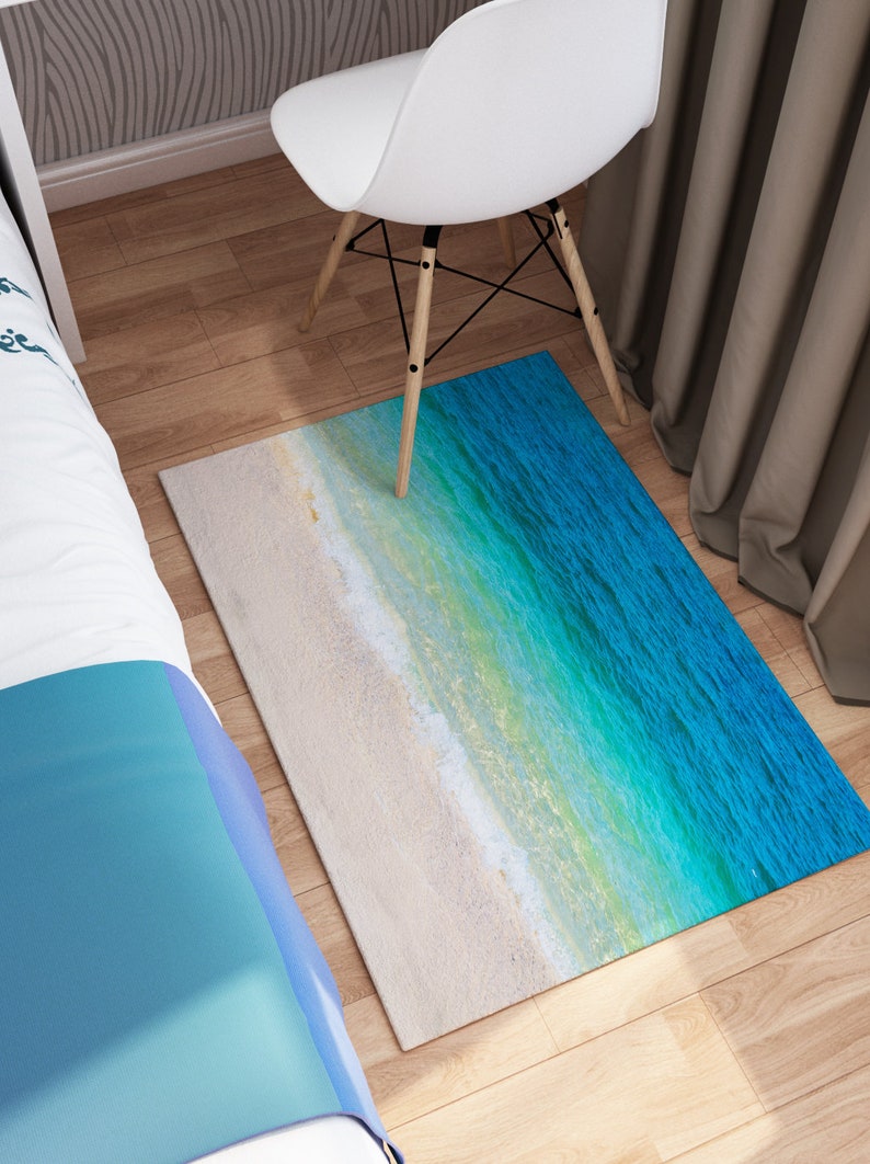 May include: Rectangular rug featuring a beach scene with white sand and turquoise water. The rug's design transitions from a sandy beige to shades of blue and green, creating a coastal aesthetic. The rug is placed on a wooden floor.