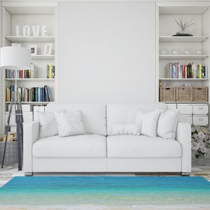 May include: A white sofa with several white pillows sits on a light-colored wooden floor. A blue and turquoise rug resembling a beach scene is in front of the sofa. White bookshelves and a floor lamp are on either side of the sofa.