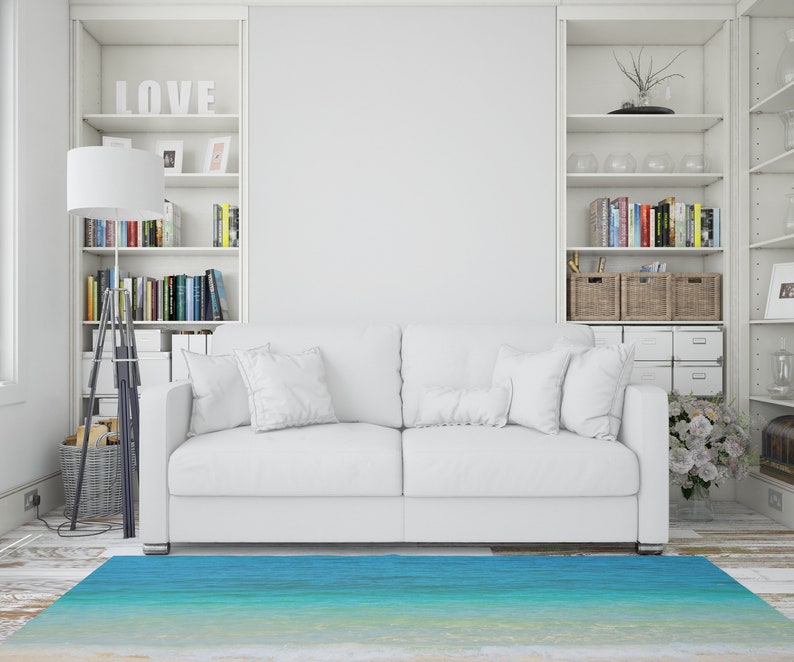 May include: A white sofa with several white pillows sits on a light-colored wooden floor. A blue and turquoise rug resembling a beach scene is in front of the sofa. White bookshelves and a floor lamp are on either side of the sofa.