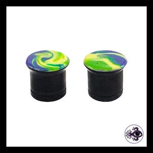 May include: Two black ear plugs with colorful, swirled designs. The tops of the plugs feature a mix of yellow, green, and blue, creating a marbled effect. The plugs are cylindrical with a flat, circular top.