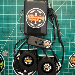 May include: A black electronic device with the text "TROOPER TRONIX" on a white and black sticker. The device has a USB cable, a switch, and two black fans. The background is a green cutting mat.