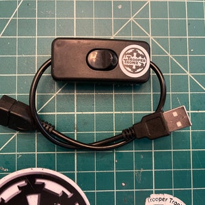 May include: A black USB switch with a black on/off button and a white sticker with the text "TROOPER TRONIX" and a logo. The switch is connected to a black USB cable.