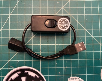 May include: A black USB switch with a black on/off button and a white sticker with the text "TROOPER TRONIX" and a logo. The switch is connected to a black USB cable.