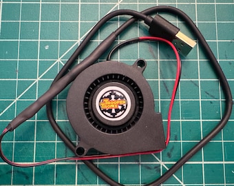 May include: A black computer fan with a USB connector and red and black wires. The fan has the text "TROOPER TRONX" on the center. The fan is on a green cutting mat.