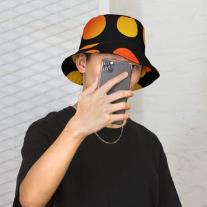 Puede incluir: Un sombrero de pescador con base negra y un patrón repetitivo de grandes puntos circulares con degradado naranja y amarillo. El sombrero tiene un ala inclinada hacia abajo y una copa plana, confeccionado en tela con un estampado uniforme.