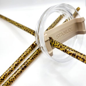May include: Two leopard print straws with a beige straw holder that says "STANLEY" on it. The straws are inserted into a clear lid.