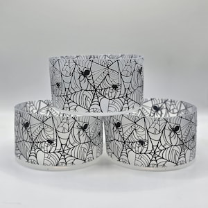 Puede incluir: Tres portavelas de vidrio esmerilado con un diseño de telaraña y araña negro. Los portavelas cilíndricos están apilados, creando una exhibición con temática de Halloween. El diseño es un patrón repetido de telarañas y arañas.