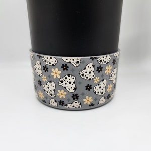 May include: A black travel mug with a gray silicone sleeve. The sleeve has a repeating pattern of white and black floral designs on a gray background.