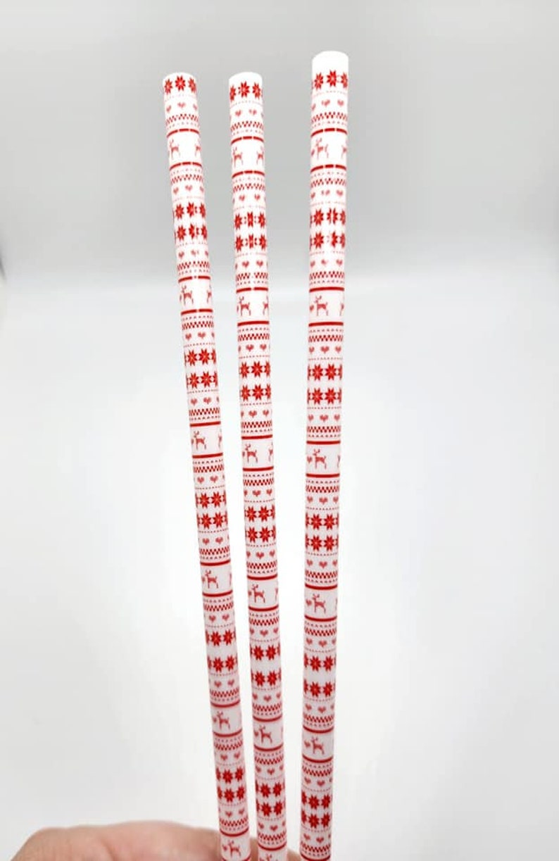 May include: Three white drinking straws with a red and white Christmas pattern. The pattern includes snowflakes, reindeer, and a geometric design.