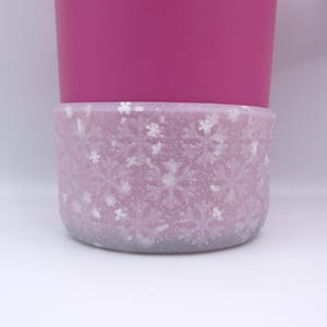 May include: A pink reusable cup with a white snowflake patterned silicone sleeve.