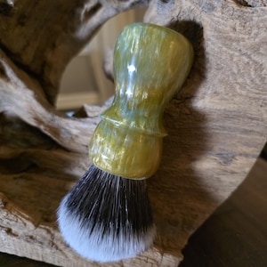 Puede incluir: Una brocha de afeitar con un mango brillante y jaspeado que presenta remolinos de color verde oliva, amarillo y blanco. El cabezal de la brocha consta de cerdas densas y oscuras que terminan en puntas blancas, creando un contraste con el mango.