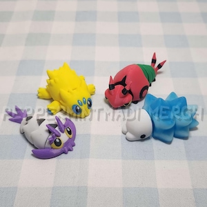 Puede incluir: Cuatro pequeñas y coloridas figuras de criaturas estilizadas. Una es amarilla con ojos azules, otra roja y verde, una tercera blanca y morada, y la última azul y blanca. Las figuras están sobre una superficie a cuadros.