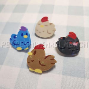 May include: Four colorful chicken-shaped pins. One is blue with a purple comb, another is beige with a red comb, a third is brown with a red comb, and the last is black with a red comb. The pins are on a checkered background.