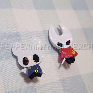 以下が含まれることがあります： 2つのキャラクター型のピン。1つは青と黒の衣装を着た白、もう1つは赤いマントを着た白です。どちらも黒い目とホログラフィックなアクセントがあります。ピンは青と白のチェック柄の表面にあります。