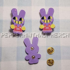May include: Three purple bunny pins with yellow and black facial features and red overalls. Two gold pin backs are also included. The text "PEPERMINTJADI MERCH" is printed on the bottom of the image.