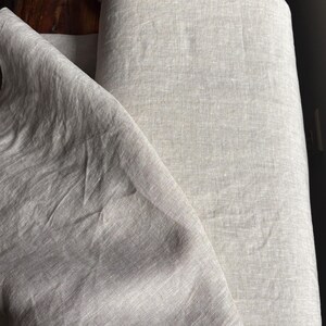 May include: A roll of natural beige linen fabric with a visible woven texture. The fabric is partially unrolled, showing soft folds and creases across the surface. The material has a neutral, earthy tone with subtle variations in the weave.