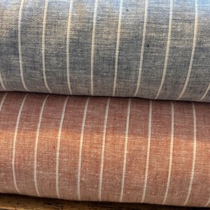 May include: Close-up of two rolls of fabric. The top roll is blue with vertical white stripes. The bottom roll is a reddish-brown colour with vertical white stripes. The fabric appears to be linen or a similar woven material.