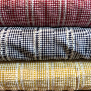 May include: Three folded bolts of seersucker fabric stacked vertically. The top bolt is red and white gingham, the middle is navy blue and white gingham, and the bottom is yellow and white gingham. Each features vertical white stripes.