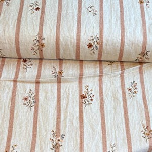 May include: A bolt of off-white fabric featuring vertical terracotta-coloured stripes and a repeating pattern of small, delicate floral sprigs in shades of brown, orange, and muted green scattered across the surface of the textile material.