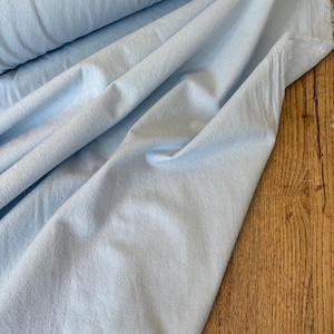 May include: A bolt of light blue fabric with a soft, matte texture. The material is draped in loose, flowing folds, revealing a slightly uneven, natural weave. The edges of the textile appear raw and unfinished, showing a subtle, fibrous fringe.