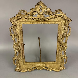 May include: Ornate gold-colored picture frame with intricate scrollwork and a decorative top. The frame has a rectangular opening and a stand on the back. The frame is made of metal and has a vintage aesthetic.