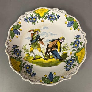 May include: A white ceramic plate with a scalloped edge, featuring a hand-painted scene of two figures in period clothing. The plate is decorated with blue floral and yellow accents.