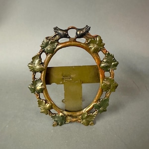 May include: An oval-shaped picture frame with a decorative design. The frame is made of metal with a gold and bronze finish. The frame is adorned with green leaves and two small birds at the top. The frame has a stand on the back.