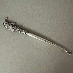 May include: A silver hook with a detailed face design on the handle. The hook is used for pulling yarn or thread through a loop.