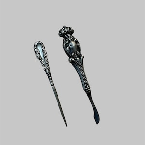 May include: Two antique silver serving utensils. One is a long, slender pick with an ornate handle. The other is a butter knife with a decorative handle topped with a crown design. Both have a vintage aesthetic.