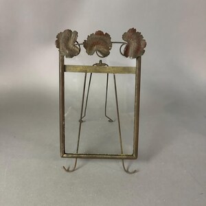 May include: An antique bronze-toned picture frame with a rectangular glass pane. The frame has a decorative top with three leaf-shaped accents. It stands on four thin legs and two curved feet.