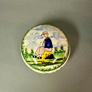 May include: A round, ceramic trinket box with a painted scene of a person sitting on a yellow rock. The person wears a blue cape and striped pants. The background features a blue sky with clouds and green trees.