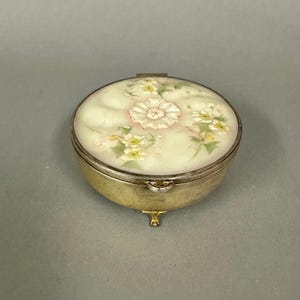 May include: A small, round, antique pillbox with a floral design. The lid features a cream-colored background with a central pink and white flower and smaller yellow flowers. The box has a silver rim and a gold-colored base with small feet.