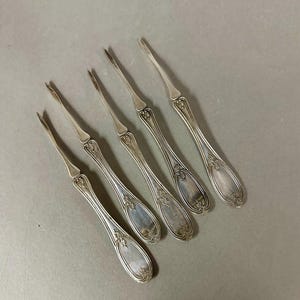 May include: Five silver-colored, antique forks with ornate floral designs on the handles. The forks have two tines and are arranged diagonally on a gray surface. The forks are likely for serving appetizers or desserts.