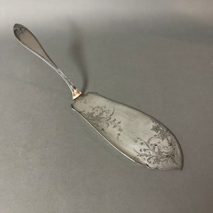 May include: Silver-toned serving utensil with a long, slender handle and a wide, curved blade. The blade features intricate floral and starburst engravings. The handle has a heart-shaped design at the end. A decorative piece for serving.