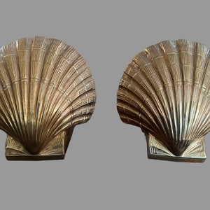 May include: A pair of gold-colored metal bookends shaped like seashells. The bookends have a detailed design of a seashell with a ribbed texture.