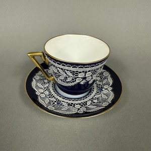 May include: A blue and white porcelain teacup and saucer set with a delicate lace design. The cup has a gold rim and a gold handle. The saucer has a gold rim and a gold band around the edge.