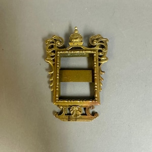 May include: A rectangular brass frame featuring ornate scrollwork along the sides, a small decorative finial at the top, and a horizontal bar across the center. The metal has a warm, metallic gold tone with intricate relief detailing throughout the piece.