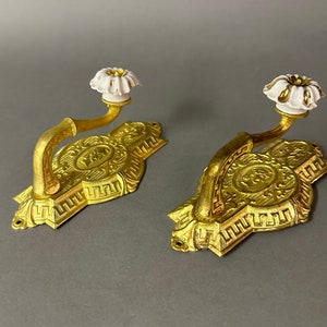 May include: Two ornate gold-toned wall hooks with white ceramic floral finials. Each hook features a detailed, embossed design with a central oval motif.