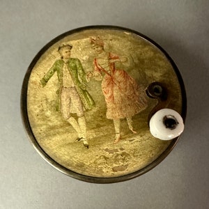 May include: A vintage music box with a hand-painted scene of a couple dancing. The music box is made of metal and has a white porcelain crank.
