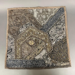 May include: Square decorative pillow with a patchwork design. The pillow features a variety of fabrics in shades of gray, brown, and gold, with intricate embroidery and beadwork. The edges are trimmed with a neutral-colored fabric.