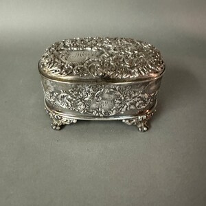 May include: Ornate silver jewelry box with floral and scroll detailing. The box is rectangular with rounded edges and rests on four small feet. The lid and sides are covered in intricate embossed designs, with a monogram on the lid.
