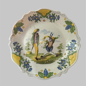 May include: A decorative ceramic plate with a scalloped edge. The plate features a hand-painted scene of two figures in period clothing, set against a green and yellow landscape. Blue floral accents adorn the rim, adding a touch of elegance.
