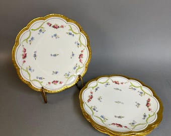 May include: A set of white porcelain plates with scalloped edges and gold-painted rims. The plates feature a delicate floral pattern of small pink roses and tiny blue flowers, connected by light green leafy garlands around the border.