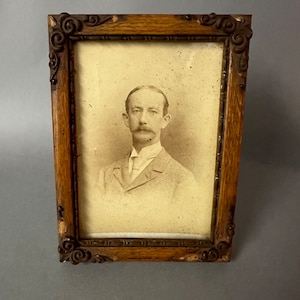 May include: A vintage wooden picture frame with ornate carvings and a faded photograph of a man with a mustache wearing a suit and tie.
