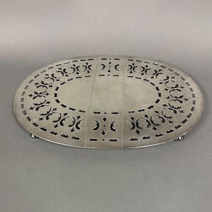 May include: Silver oval-shaped trivet with a decorative cut-out design. The trivet has three legs and is made of a shiny metal.