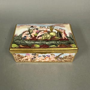 May include: A rectangular, antique porcelain box with a gold trim. The lid features a painted scene of cherubs and a landscape. The sides of the box also have cherub designs. The box is likely used for jewelry or small keepsakes.