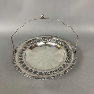 May include: A silver serving tray with a decorative border and a handle. The tray is round and has a slightly raised edge.