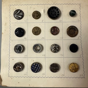 May include: A collection of 15 vintage buttons displayed on a white card with handwritten numbers indicating their size. The buttons are various colors, shapes, and materials, including metal, plastic, and glass.